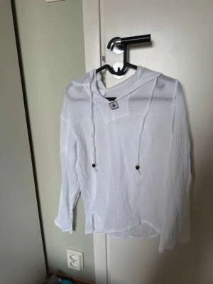 Vit tunn långärmad blus med huva - Beach cover up från Thailand. En vit, tunn och luftig tröja i 100% bomull med huva och långa ärmar. Har snörning vid halsen och är aldrig använd❤️skulle säga att den är mindre i storlek?