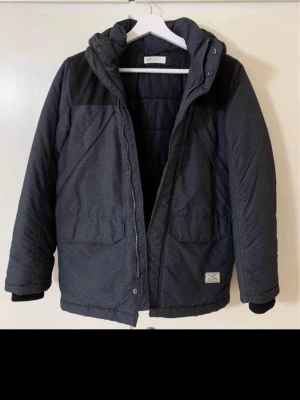 Mörkgrå pufferjacka från H&M - Säljer en mörkgrå pufferjacka från H&M med huva och stora fickor framtill. Jackan har svarta detaljer vid axlarna och dragkedja samt tryckknappar. Perfekt för kyliga dagar och har en schysst, enkel look.