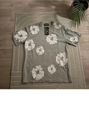 Grå blommig t-shirt från Denim Tears - Säljer en grå t-shirt från Denim Tears med stora vita blommor tryckta över hela plagget. T-shirten har rund hals och korta ärmar. Materialet är 100% bomull och känns mjukt mot huden. Perfekt för dig som gillar streetwear med unik design.