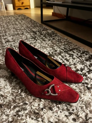 Röda pumps från Marco Tozzi - Snygga röda pumps från Marco Tozzi med spetsig tå och låg klack. Skorna är gjorda i mocka och har en dekorativ metallspänne framtill. 