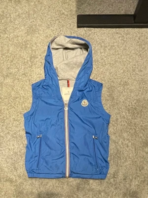 Blå Moncler dunväst med huva - Snygg blå dunväst från Moncler med grå huva och dragkedja framtill. Västen har två fickor med dragkedja och Moncler-logga på bröstet. Insidan är grå och materialet känns lätt och sportigt. Perfekt för lager-på-lager och streetstyle.