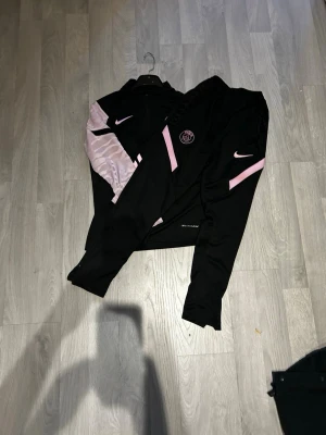 Svart och rosa PSG Nike träningsset - Säljer ett fett träningsset från Nike x PSG med långärmad tröja och byxor. Setet är svart med rosa detaljer och PSG-logga på både tröja och byxa. Tröjan har halv dragkedja och mönstrade ärmar i ljusrosa. Materialet är polyester med Dri-Fit teknologi.
