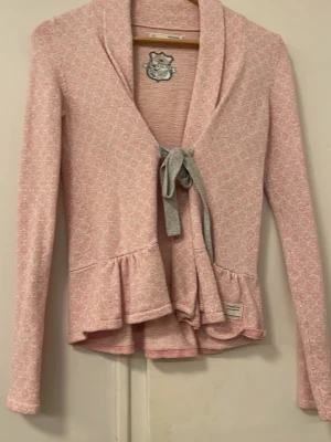 Rosa kofta - Supersöt rosa kofta från Odd Molly med diskret mönster och grått knytband framtill. Koftan har volangdetaljer vid nederkanten och långa ärmar. Perfekt för dig som gillar romantisk och feminin stil. Materialet känns mjukt och bekvämt mot huden.