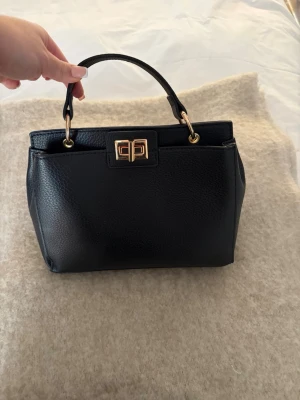 Svart handväska med gulddetaljer - Snygg svart handväska i skinn med guldfärgade metalldetalje. Väskan har flera fack inuti och ett elegant lås framtill. Perfekt storlek för att få plats med allt viktigt och har en stilren, klassisk form. Väskan är aldrig använd så i fint skick, skriv privat för fler bilder eller om du undrar något!🩷