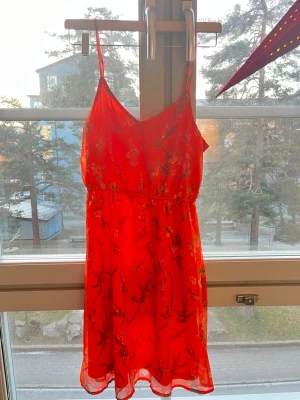 Orange blommig kortklänning med smala axelband - Säljer en röd kortklänning med smala axelband och blommigt mönster. Klänningen har en lätt och luftig känsla, med markerad midja och v-ringad hals. Perfekt för varma dagar och sommarhäng. Materialet känns som polyester.