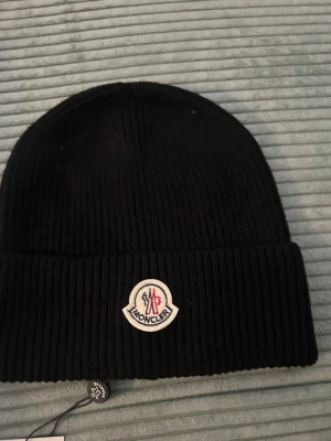 Svart Moncler mössa i ribbstickad ull - Svart ribbstickad mössa från Moncler med klassisk uppvikt kant och broderad logotyp framtill. Mössan är tillverkad i mjuk ull och har en stilren look som passar perfekt när det är kallt ute. Taggen sitter kvar.