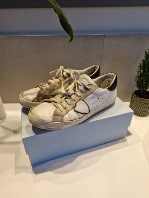 Phillipe model vita sneakers - Säljer ett par vita sneakers från Phillipe model med svarta detaljer på hälen och gråa inslag på sidorna. Skorna har snörning och rund tå, tillverkade i skinn och mocka. Perfekta för dig som gillar stilrena och lyxiga sneakers med cool design.