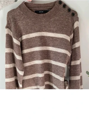 Brun randig stickad tröja Vero Moda - Mysig brun stickad tröja från Vero Moda med beige/ vita ränder och fyra knappar på ena axeln. Tröjan har rund halsringning, lång ärm och avslappnad passform. Perfekt för höst och vinter när du vill vara både varm och snygg.