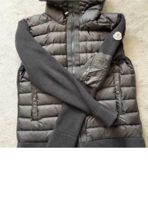 Svart cardigan moncler  - Säljer en svart pufferjacka från Moncler med stickade ärmar och quiltad kropp. Jackan har dragkedja framtill, fickor med knappar och en extra ficka med dragkedja på ärmen. Moncler-logga på vänster ärm. Perfekt för dig som vill ha en snygg och varm jacka med street vibe.