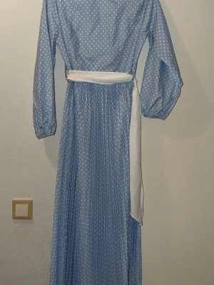 Blå prickig långklänning med bälte - Säljer en blå långklänning med vita prickar och ett vitt bälte i midjan. Klänningen har långärmade puffärmar och ett plisserat fall som ger en snygg siluett. Perfekt för dig som gillar retrostil och vill sticka ut med ett klassiskt mönster.