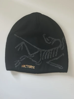Svart mössa från Arc'teryx - Svart mössa från Arc'teryx med grått grafiskt mönster på framsidan och broderad logga i guld. Mössan är enkel och stilren, perfekt för kalla dagar. Materialet känns mjukt och stretchigt, vilket gör den bekväm att bära.