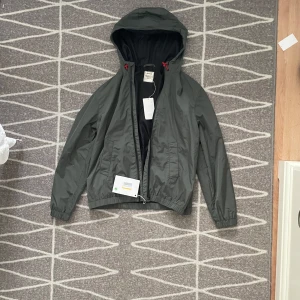 Grå vindjacka med huva från Zadig & Voltaire - Snygg mörkgrön vindjacka från Zadig & Voltaire med huva och röda dragskor. Jackan har dragkedja framtill, elastiska muddar och två fickor. Perfekt för blåsiga dagar och enkel att matcha med olika outfits.