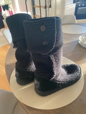 Svarta stickade UGGs med knappar - Säljer ett par svarta UGG boots i stickad ull med två dekorativa knappar på skaftet. Skorna har platt sula och rund tå, samt UGG-logga baktill. Perfekt för dig som gillar mysiga och varma skor med cool design.