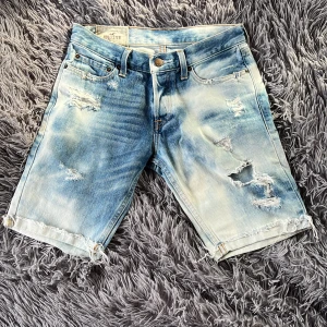 Blå slitna jeansshorts från Hollister - Säljer ett par blå jeansshorts från Hollister med slitna detaljer och fransiga benslut. Shortsen har fem fickor, knappar framtill samt coolt tvättad look med ljusa och mörka partier. Perfekt för dig som gillar streetstyle. 