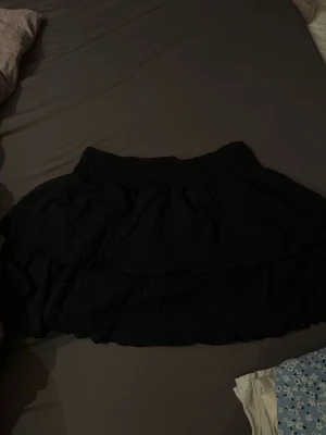 Svart volangkjol från Gina Tricot - Säljer en svart kortkjol från Gina Tricot i storlek L. Kjolen har bred resår i midjan och flera lager med volanger som ger en snygg och luftig look. Materialet är 100% bomull och kjolen är perfekt för dig som gillar enkel men trendig stil.