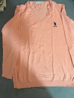 Ljusrosa v-ringad tröja U.S. Polo.  - Snygg ljusrosa stickad tröja från U.S. Polo . med v-ringad hals och broderad marinblå logga på bröstet. Tröjan har långa ärmar och ribbade muddar vid ärmslut och nederkant. Perfekt för dig som gillar klassisk och stilren design. Jätte fin och stilig och har ingens kada och om de finns något skada på den så kan du returnera direkt tillbaka. 
