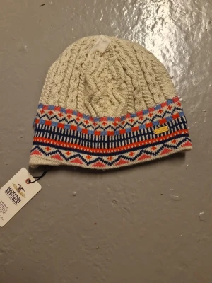 Stickad mössa från Hampton Republic - Snygg stickad mössa från Hampton Republic i ljus beige med flätstickat mönster upptill och färgglada geometriska detaljer i blått, rött och rosa nedtill. Perfekt för kalla dagar och har en liten guldfärgad logga på sidan. För lite smallare huvuden