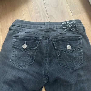True Religion Becca Bootcut Jeans gråa - Säljer ett par gråa jeans från True Religion, modell Becca Mid Rise Bootcut. Jeansen har snygga silverfärgade knappar och detaljerade bakfickor med broderad logga. Klassisk femficksdesign och kontrastsömmar ger en cool look. Materialet är mjukt jeans-tyg. Jeansen har en bootcut passform och sitter tightare runt låren men utsvängda längs vaderna. Lite korta på mig som är ca 175cm lång. 