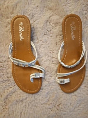Vita sandaler med strass från Bonita - Säljer ett par snygga sandaler från Bonita med vita remmar och glittrande strassdetaljer. Sandalerna har platt sula och öppen tå, med dekorativa fyrkantiga strass på remmarna. Perfekta för sommarens varma dagar och ger en fräsch look.