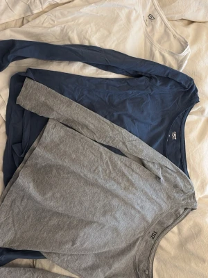 3-pack långärmade toppar från 157 - Säljer tre stilrena långärmade toppar från 157 i storlek XS. En är vit, en marinblå och en ljusgrå, alla med rund halsringning och enkel design. Perfekta att matcha med jeans eller kjol. Materialet är mjuk bomull med lite stretch för skön passform.