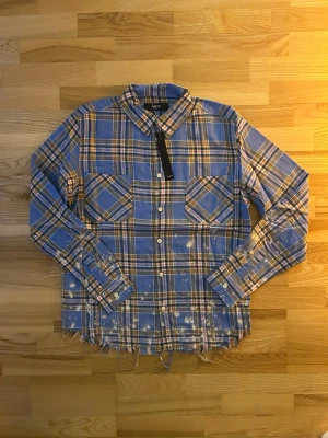 Blå rutig skjorta från Amiri - Säljer en blå rutig skjorta från Amiri med stora vita bokstäver på ryggen. Skjortan har två bröstfickor, knappar framtill och fransad nederkant. Materialet är mjuk bomull och färgerna är blå, brun och vit med inslag av rött. Perfekt för dig som gillar streetwear och detaljer.