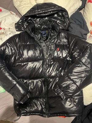 Svart pufferjacka från Polo Ralph Lauren - Svart glansig pufferjacka från Polo Ralph Lauren med huva och dragkedja framtill. Jackan har ett rött broderat Polo-logo på bröstet och flera fickor med dragkedja. Materialet är syntet och jackan är quiltad för extra värme. Perfekt för kalla dagar.