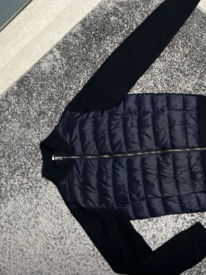 Mörkblå cardigan - Säljer en  mörkblå cardigan. Framsidan är quiltad och har en hel dragkedja. Jackan har en cool mix av material med blankt syntet framtill och mjukt stickat tyg på ärmar och baksida. Perfekt för dig som vill ha en unik look. Märket heter Maglia studios och orginalpris för den är 2000kr.