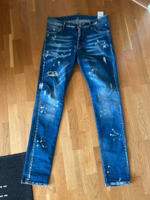  DSQUARED2 jeans med slitningar - Säljer ett par blå jeans från DSQUARED2 med coola slitningar och färgstänk på både fram- och baksida. Jeansen har klassisk femficksdesign, rak passform och är tillverkade i bomull. Märkespatch bak i midjan och snygga detaljer som ger en edgy vibe.