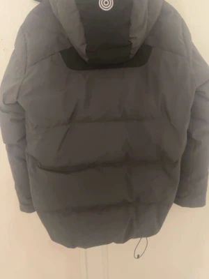 Mörkgrå dunjacka från Sail Racing - Säljer en mörkgrå dunjacka från Sail Racing med svarta detaljer och röd insida. Jackan har huva, två dragkedjeförsedda bröstfickor och justerbar nederkant med snörning. Perfekt för kalla dagar och har en clean, sportig look. Helt oanvänd