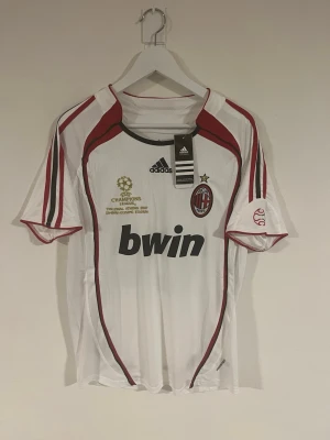 AC Milan 2006/07 Kaka' 22 CL tröja - Säljer en officiell AC Milan Champions League 2007 tröja med Kaka' och nummer 22 på ryggen. Tröjan är vit med röda och svarta detaljer, Adidas-logga och Bwin sponsortryck. Broderat Champions League-märke och ACM-emblem på bröstet. Perfekt för fotbollsfans!