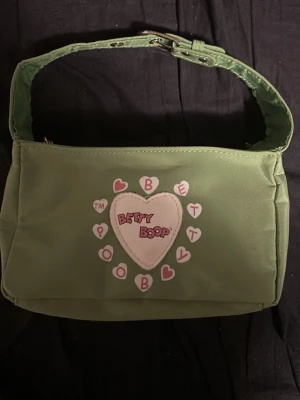 Grön Betty Boop axelväska - Säljer en grön axelväska från Betty Boop med justerbart axelband och dragkedja. Väskan har ett hjärtmotiv framtill med Betty Boop-logga och små rosa hjärtan runtom. 