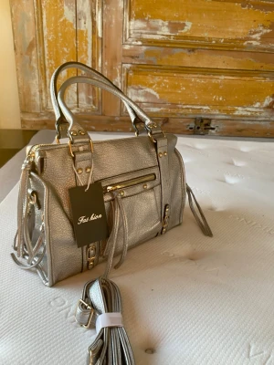 Silvrig handväska med detaljer - Silvrig handväska till orginalpris från Fashion med guldfärgade metalldetaljer och flera dragkedjor. Rymmer precis en MacBook.