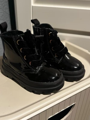  Barn lackade kängor med snörning svarta de 21/22 - Säljer ett par svarta kängor från H&M med glansig lackad yta och grov sula. Skorna har rund tå, snörning med breda svarta band och guldfärgade öljetter. Perfekt för dig som gillar chunky boots med attityd.