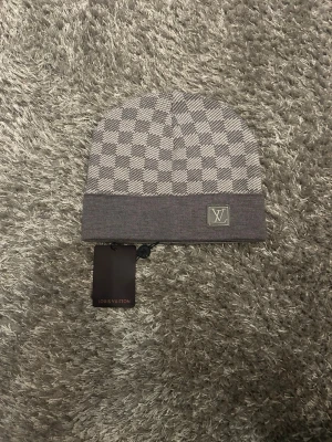 Grå Louis Vuitton mössa - Snygg grå mössa från Louis Vuitton med klassiskt rutigt mönster och LV-logga framtill. Mössan har bred uppvikt kant och är stickad i mjukt material som känns lyxigt. Perfekt accessoar för dig som gillar designer och vill ha något som sticker ut.