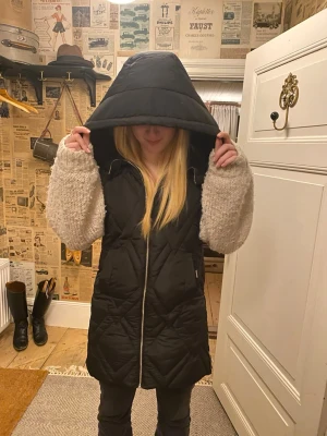 Svart lång quiltad dunväst med huva - Säljer en svart lång dunväst med stor huva och quiltat mönster. Västen har dragkedja framtill och dragkedjor på sidorna. Västen kommer inte till andvändning och är helt ny med lappar kvar på.💕