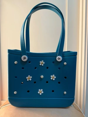 Blå Eva väska med blommor - Säljer en blå väskamed två rejäla handtag. Väskan är dekorerad med vita blommor med gul mitt och pärldetaljer på framsidan. Materialet är syntet och väskan har ett lekfullt och unikt utseende med hålmönster. Perfekt för dig som gillar färg och detaljer!