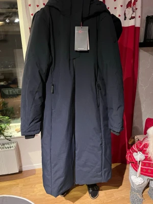 Mörkblå lång ridkappa från Covalliero - Säljer en mörkblå lång dunjacka från Covalliero med huva och dragkedja. Jackan har två sidofickor med dragkedja och stilren design. Perfekt för kalla dagar och har en snygg, enkel look som funkar till många outfits. Är en ridkappa köpt för 1699 men säljes för 1200! 