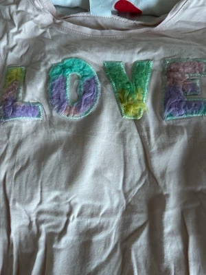 Ljusrosa t-shirt med LOVE-tryck Lindex - Söt ljusrosa t-shirt från Lindex med färgglada fluffiga bokstäver som bildar ordet LOVE på bröstet. T-shirten är ärmlös och har en knytning i sidan för extra detalj. Perfekt för dig som gillar pastell och mjuka material.