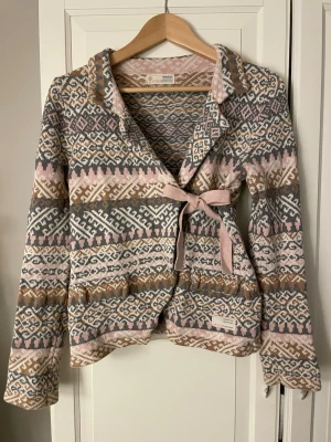 Mönstrad kofta Odd Molly med knytband - Supersnygg kofta från Odd Molly i stickad bomull med grått, beige, brunt och ljusrosa mönster. Koftan har långa ärmar, krage och ett dekorativt knytband i midjan. Broderad Odd Molly-logga på ärmen. Perfekt för lager på lager under höst och vinter.