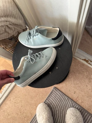 Ljusblå skor från Axel Arigato - Snygga ljusblå sneakers från Axel Arigato med vit platt sula och svart detalj på hälen. Skorna har snörning och är tillverkade i skinn för en clean och modern look. Perfekta för dig som gillar minimalistisk stil och vill ha något fräscht till vardags.