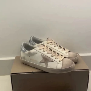 Golden Goose Superstar sneakers med glitter - Golden Goose Superstar sneakers i vitt skinn med beige mockadetaljer och snörning. Silverglittrig häl och ikonisk stjärna på sidan. Allt medföljer + låda (lånade bilder då jag aldrig använt, köpta på Plick men äktenhets bevis medföljer) skicka gärna prisförslag om du är intresserad!