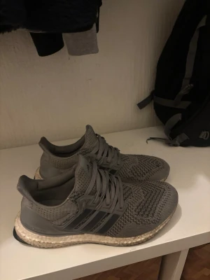 Adidas Ultraboost grå sneakers äkta - Säljer ett par grå Adidas Ultraboost sneakers med stickad ovandel och klassiska tre ränder på sidan. Skorna har snörning och en bekväm, boostad sula i vitt och beige. Perfekta för dig som gillar stilrena och sportiga skor med skön passform. Säljs inte mer någonstans!