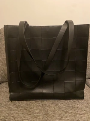 Svart rutmönstrad axelväska från ASOS - Snygg svart axelväska från ASOS med rutmönstrad design och långa axelband. Väskan är tillverkad i skinnimitation och har ett magnetlås samt en innerficka med dragkedja. Perfekt storlek för att få plats med allt du behöver under dagen.