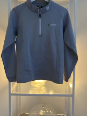 Blå fleece-tröja med half zip - Blå långärmad fleece-tröja från Stiftelsen Skånska Landskap med half zip framtill och liten logga på bröstet. Tröjan har en hög krage och är perfekt för kyliga dagar. Enkel och stilren design utan mönster eller extra detaljer. Finns en defekt på en underarmen se bild. 