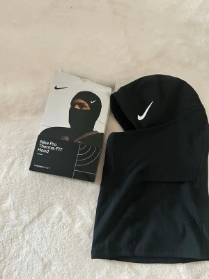 Nike Pro svart Therma-FIT huva - Nike Pro Therma-FIT Hood i svart med vit Nike-logga. Huva i mjukt och värmande syntetmaterial, designad för att ge extra skydd mot kyla. Sitter tight runt huvudet och halsen, perfekt för sport och träning utomhus. Snygg och stilren look med diskret etikett på sidan.
