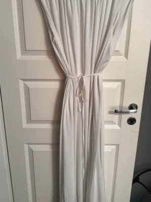 Vit klänning - Säljer en vit långklänning i mjukt och lätt material. Klänningen är ärmlös och har ett enkelt knytband i midjan som ger en snygg siluett. Den har rundad hals och ett stilrent, enfärgat utseende. Perfekt för varma dagar. Man ser ej igenom eftersom det är dubbla tyg.