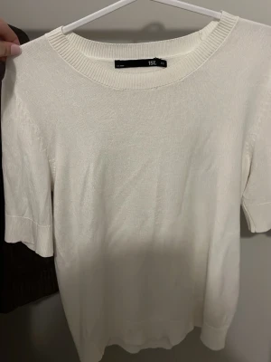 Vit kortärmad tröja från 157 - En stilren vit tröja från 157 i storlek XS. Tröjan har korta ärmar, rund halsringning med ribbad kant och är gjord i ett mjukt, tunt material som känns skönt mot huden. Perfekt till vår och sommar för en clean och enkel look.