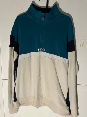 Grön och vit half zip tröja från FILA - Säljer en snygg långärmad tröja från FILA i storlek XL. Tröjan har en grön överdel, vit/beige nederdel och svarta detaljer på ärmarna. Den har en half zip dragkedja och broderad FILA-logga på bröstet. Perfekt för chill dagar och streetwear.