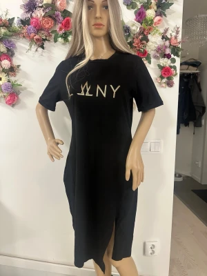 Svart t-shirtklänning från DKNY - Svart t-shirtklänning från DKNY med guldig logga framtill. Klänningen har korta ärmar, rund halsringning och en slits nertill på ena sidan. Enkel och stilren modell i mjukt material, perfekt för en avslappnad look.