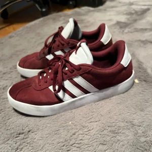 Vinröda Adidas sneakers strl 44 - Snygga vinröda Adidas vl court 3.0 sneakers  med klassiska vita ränder på sidorna och vit sula. Ovandelen är i mocka. Köptes för 3 veckor sen helt nya, säljes då dom inte passade mig 
